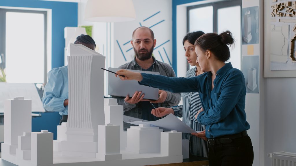 multi ethnic people working with laptop tablet building model architecture project coworkers doing teamwork design maquette construction layout with blueprints x